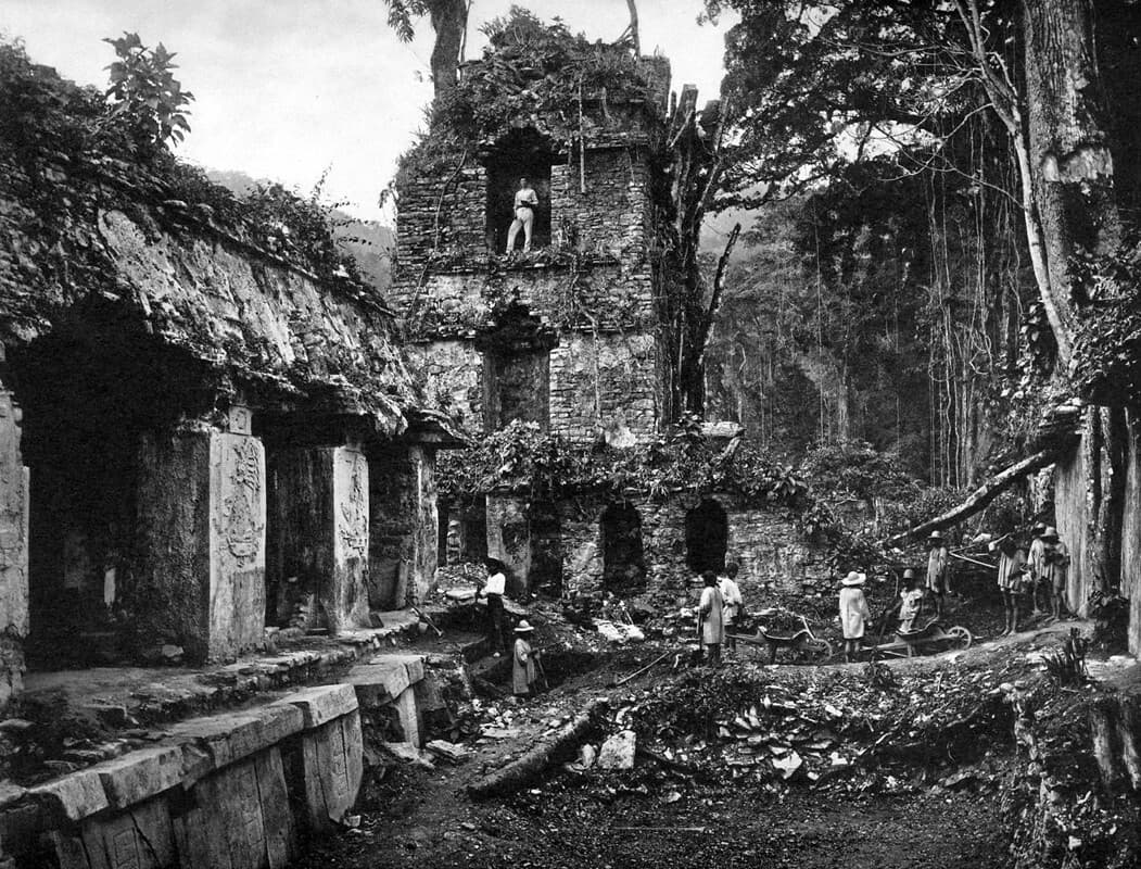 A group of people explore and work on the excavation of ancient ruins surrounded by dense jungle. The stone structures are overgrown with vegetation.