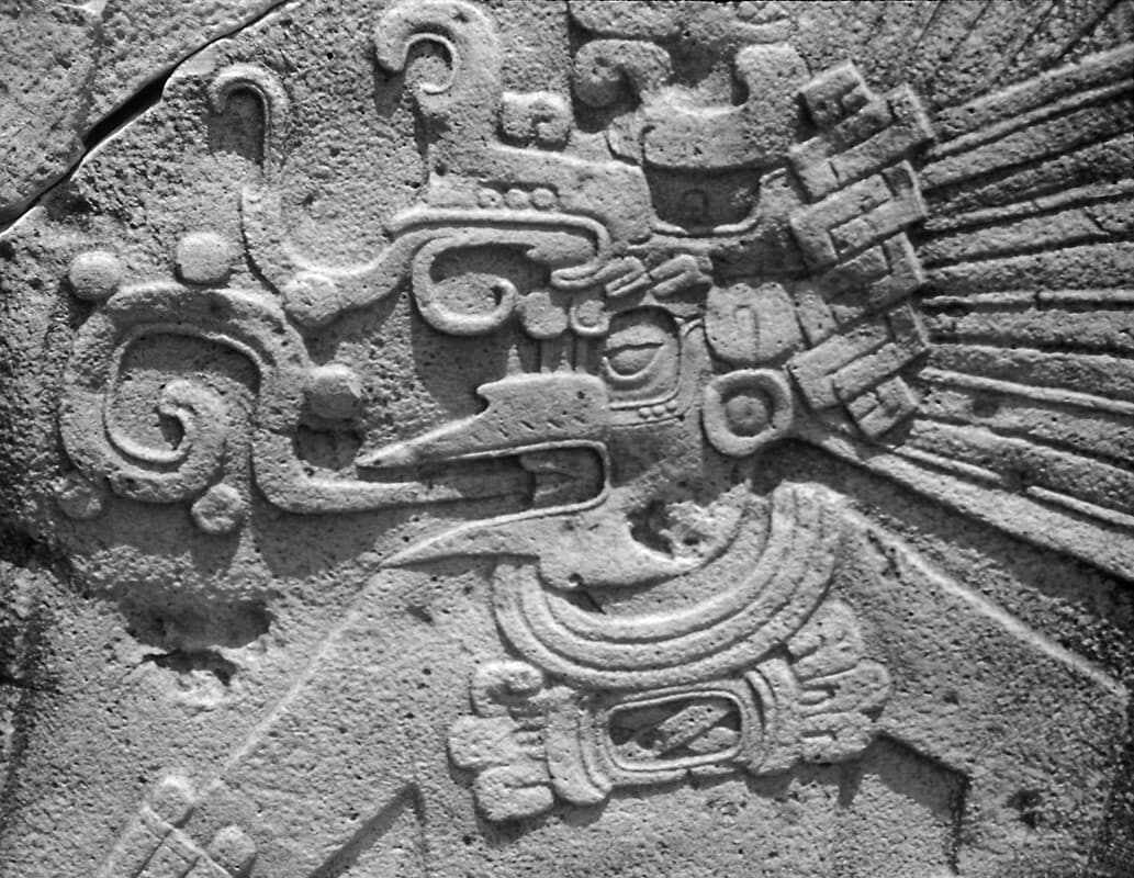 Stone carving of a Mayan figure wearing an elaborate headdress, blowing out swirling patterns. The figure's attire and intricate details indicate its cultural significance.